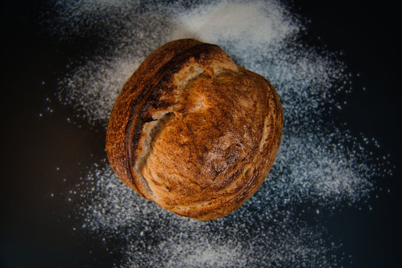 gallery-03 A top-down view of a rustic artisanal bread loaf with scattered flour, set against a dark background.